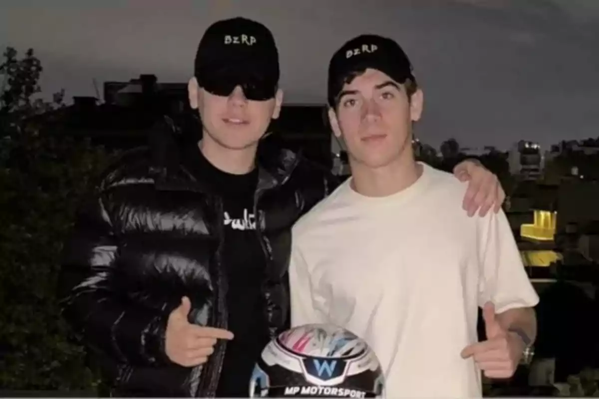 Dos personas con gorras negras posan juntas al aire libre sosteniendo un casco de carreras.