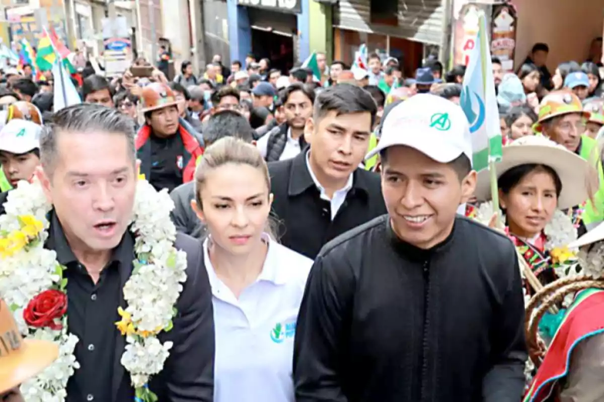 Un grupo de personas camina en una calle concurrida, algunas llevan guirnaldas de flores y una persona sostiene una bandera.