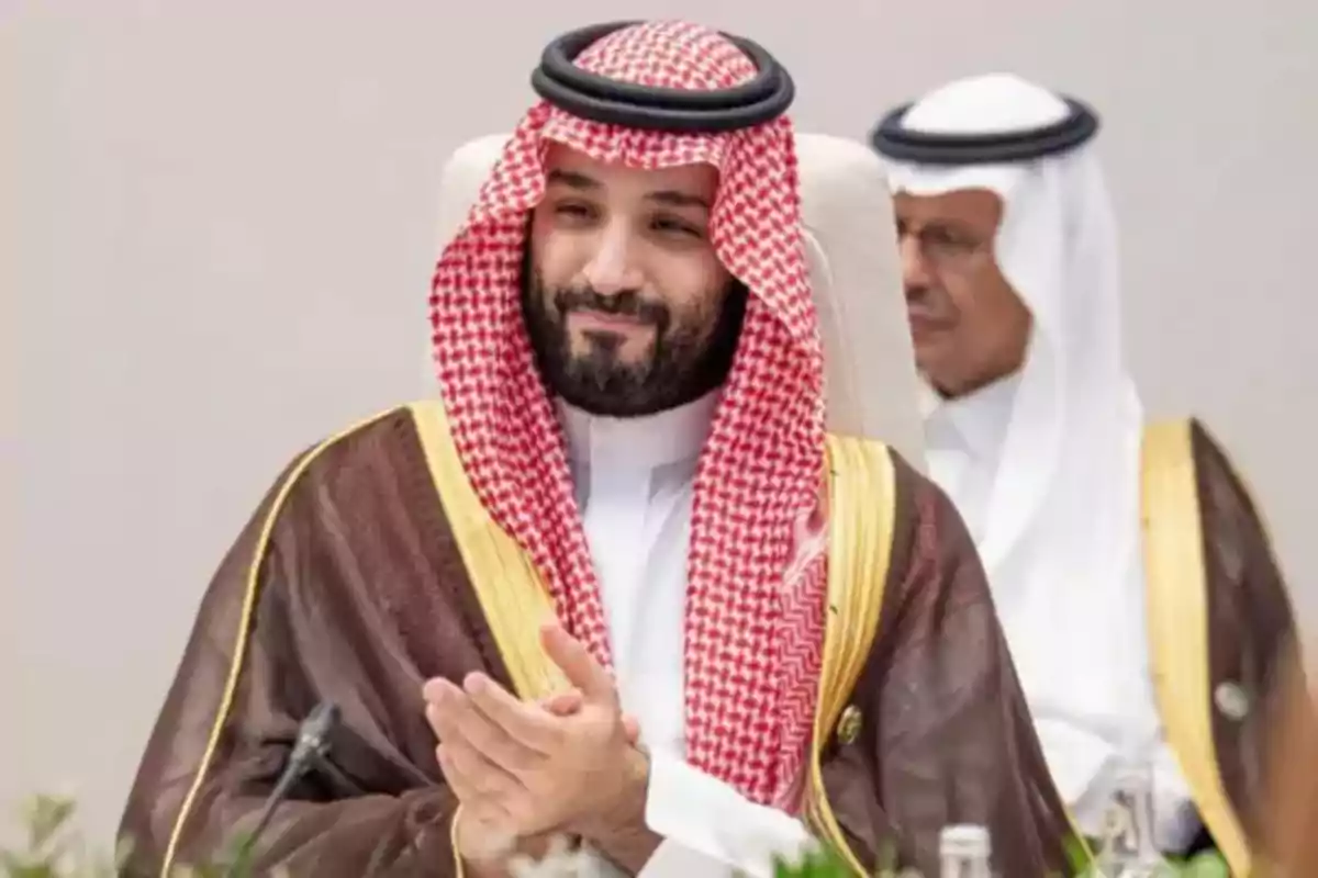A man in traditional Arab attire with a red and white headscarf is sitting and clapping at a formal event. A man in traditional Arab attire with a red and white headscarf is sitting and clapping at a formal event.