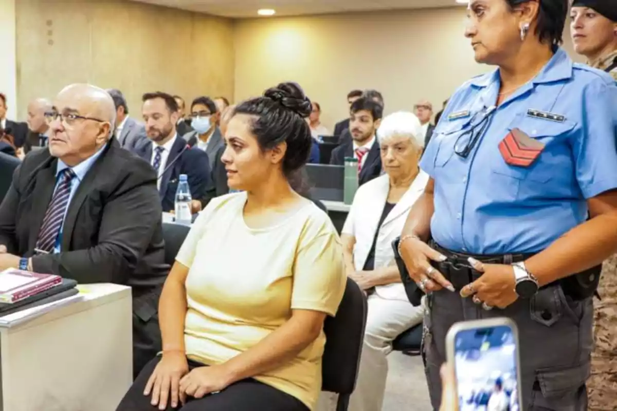 Mujer escoltada por una oficial de policía en una sala de audiencias llena de personas, algunas de ellas con traje y otras tomando notas Mujer escoltada por una oficial de policía en una sala de audiencias llena de personas, algunas de ellas con traje y otras tomando notas