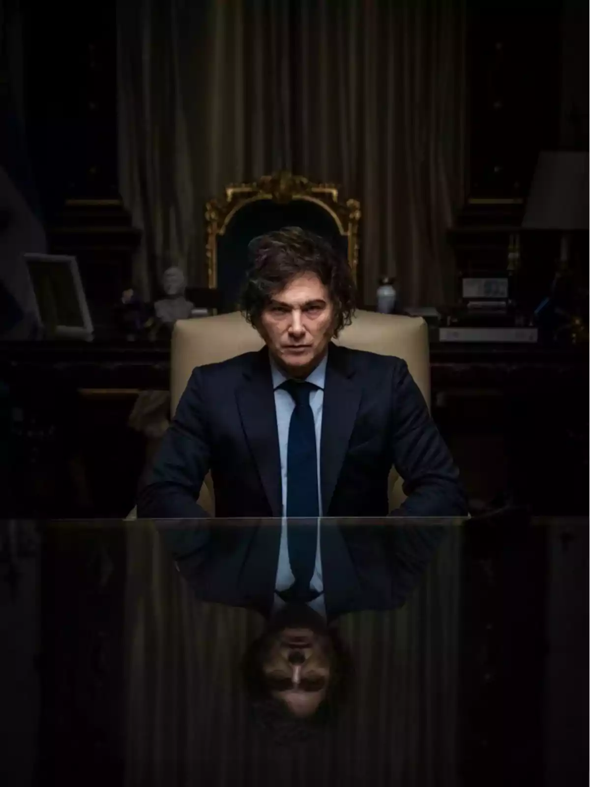 Hombre de cabello ondulado y traje oscuro sentado en un escritorio elegante con su reflejo visible en la superficie pulida de la mesa Hombre de cabello ondulado y traje oscuro sentado en un escritorio elegante con su reflejo visible en la superficie pulida de la mesa