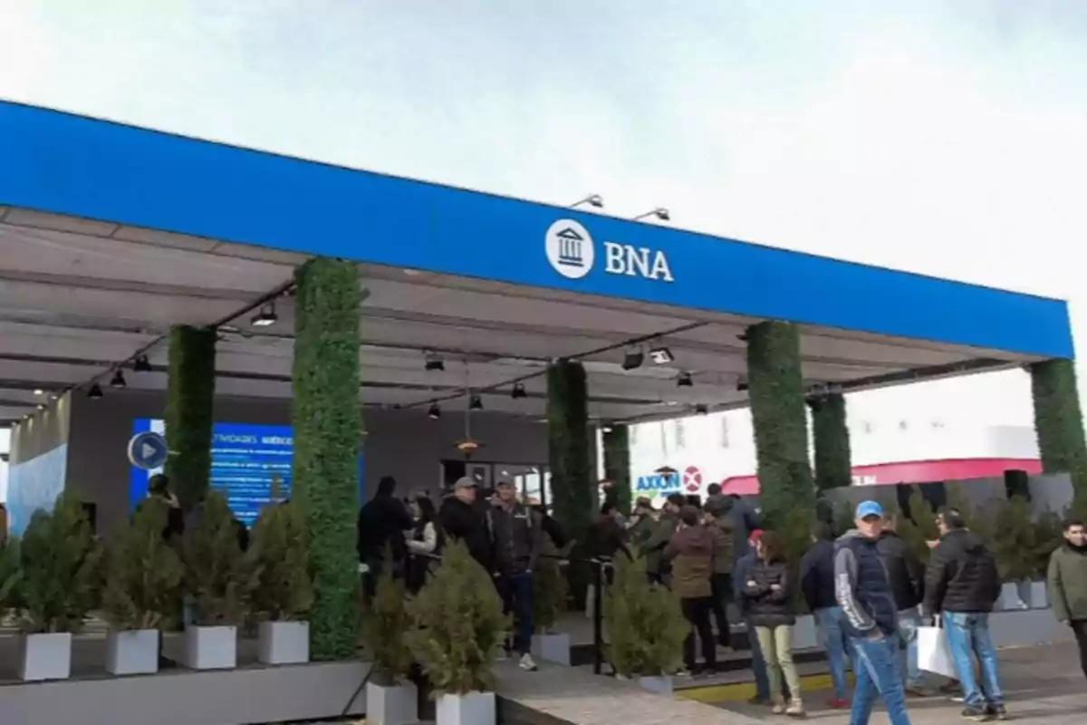 Personas reunidas en la entrada de un stand del Banco Nación decorado con plantas y un cartel azul con el logo BNA
