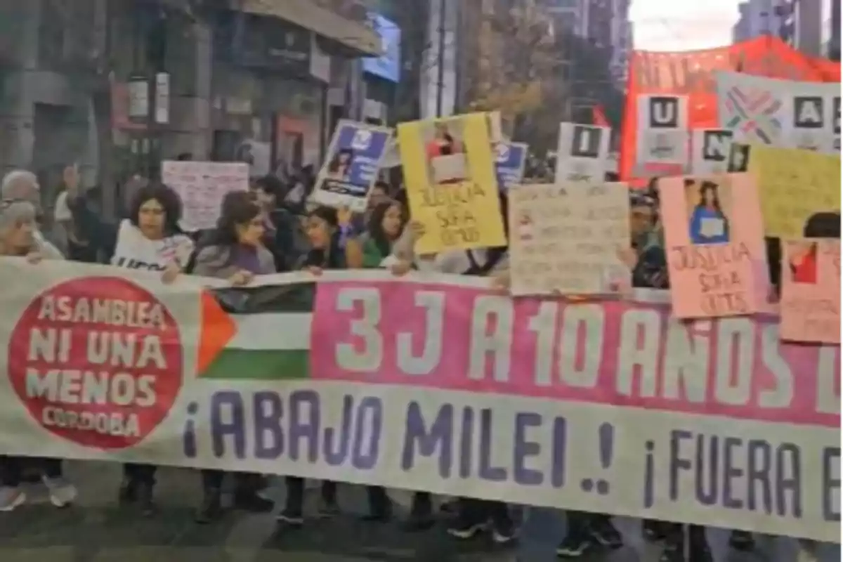 Feminist protest in the street with people holding signs and a large flag that reads "Asamblea Ni Una Menos Córdoba" and messages against Milei's government.
