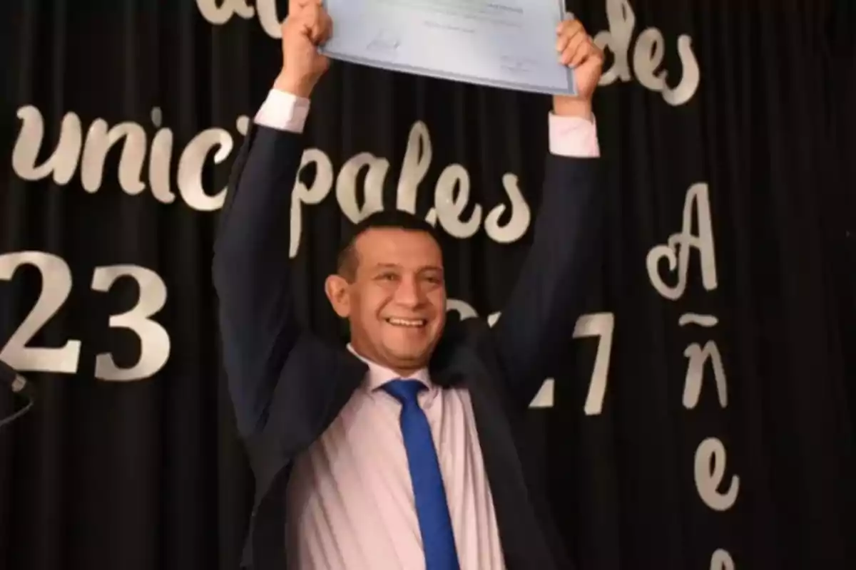 Hombre sonriente con traje y corbata azul levantando un diploma en un evento de premiación frente a un fondo negro con letras blancas decorativas Hombre sonriente con traje y corbata azul levantando un diploma en un evento de premiación frente a un fondo negro con letras blancas decorativas