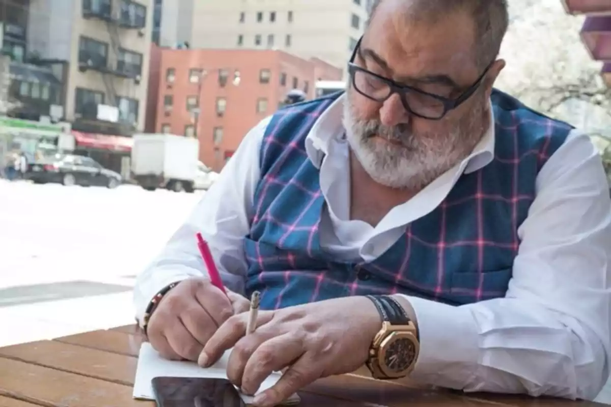 Hombre con barba y gafas escribiendo en un cuaderno mientras sostiene un cigarrillo en una mesa al aire libre en un entorno urbano.