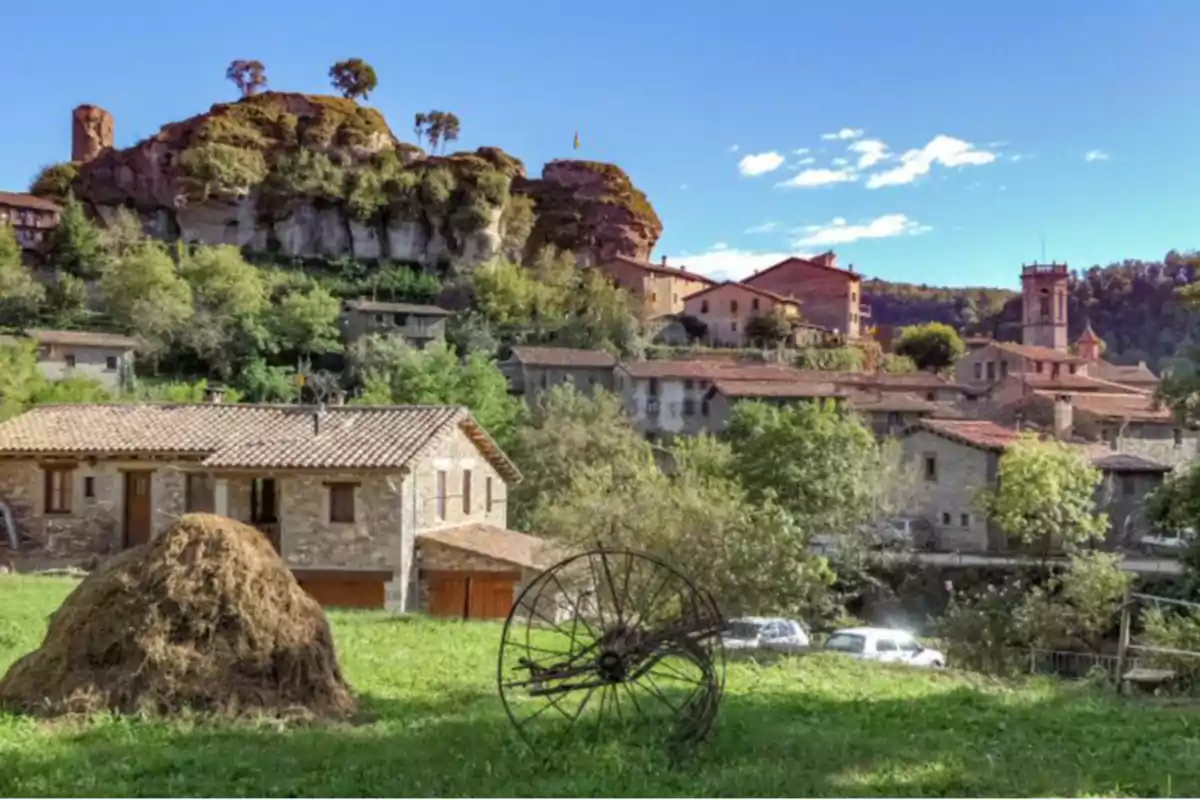 Vista de un pintoresco pueblo con casas de piedra rodeadas de vegetación y una colina rocosa al fondo bajo un cielo azul.