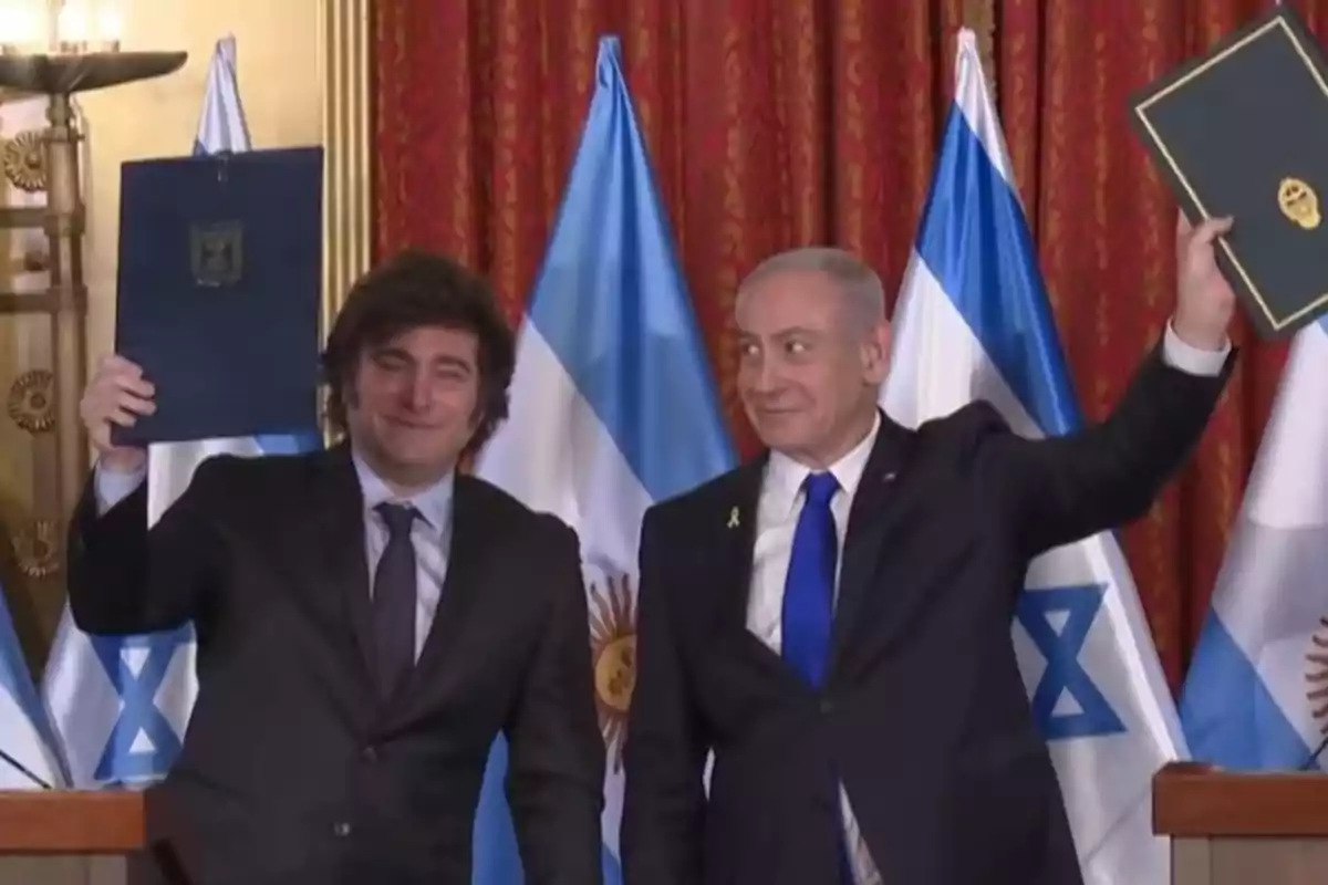 Dos hombres de traje sonríen y levantan carpetas en un acto oficial con banderas de Argentina e Israel de fondo