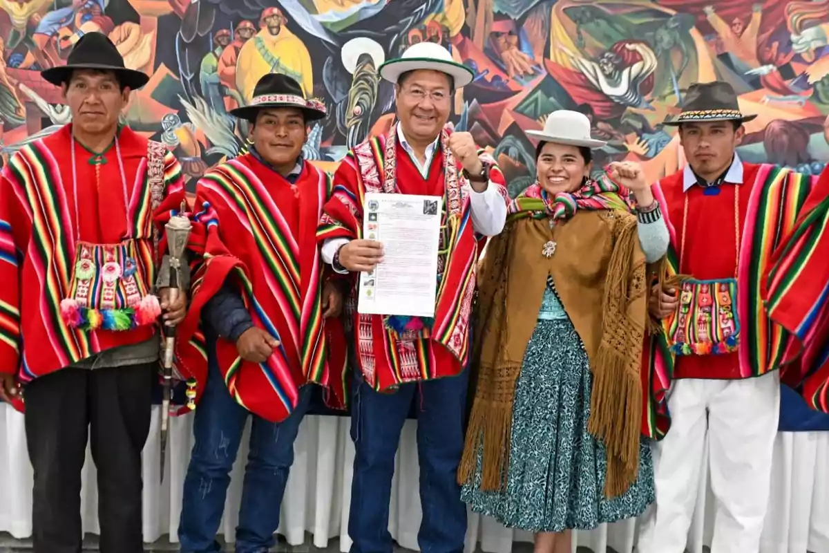 Un grupo de personas vestidas con trajes tradicionales coloridos posan frente a un mural vibrante, mostrando un documento y levantando los puños en señal de celebración.