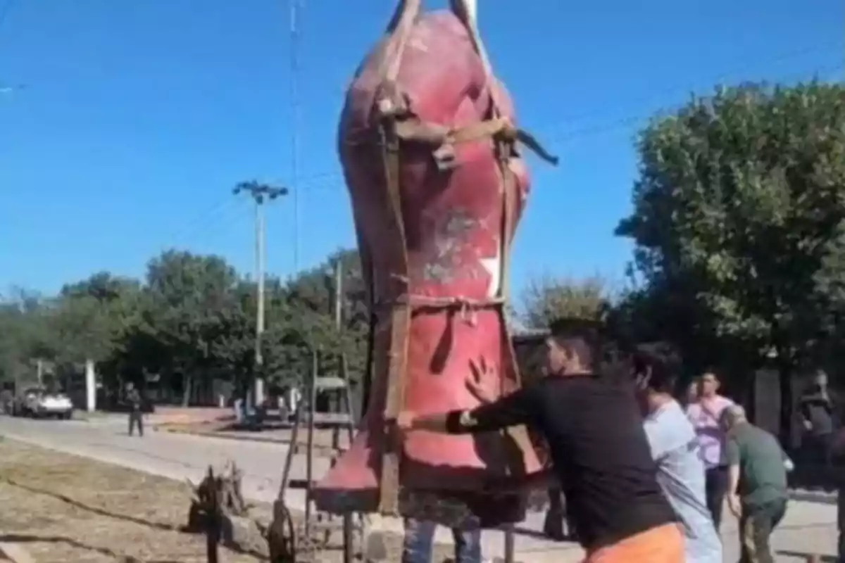 A group of people is moving a large red figure on an open-air street, surrounded by trees and light poles. A group of people is moving a large red figure on an open-air street, surrounded by trees and light poles.