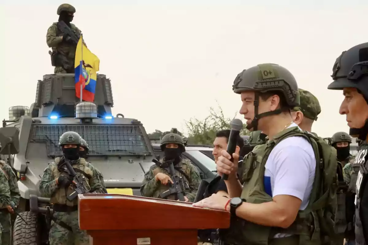 A group of military and police with tactical gear, one of them speaking into a microphone at a podium, with an armored vehicle and a flag in the background.