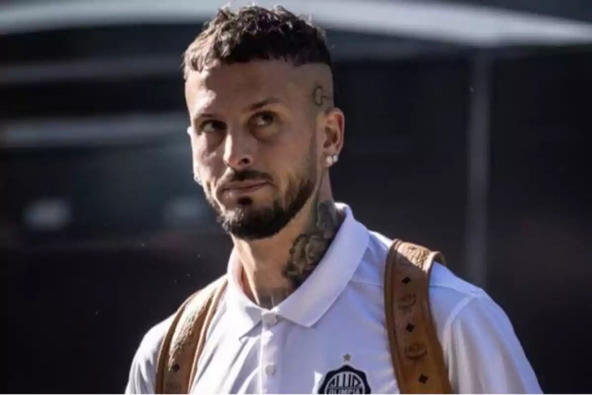 Un hombre con barba y tatuajes lleva una camiseta blanca con un logotipo y una mochila marrón.