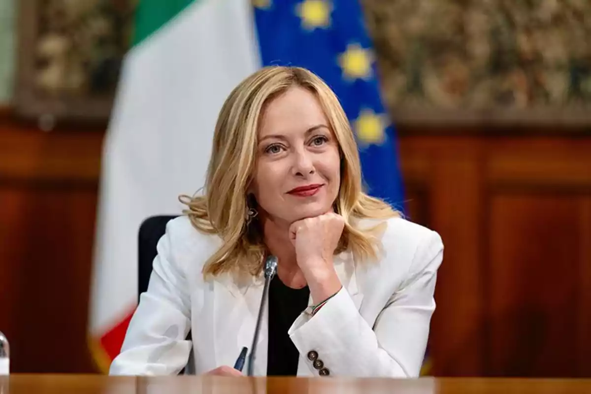 Una mujer rubia con chaqueta blanca está sentada frente a un micrófono con banderas de fondo.