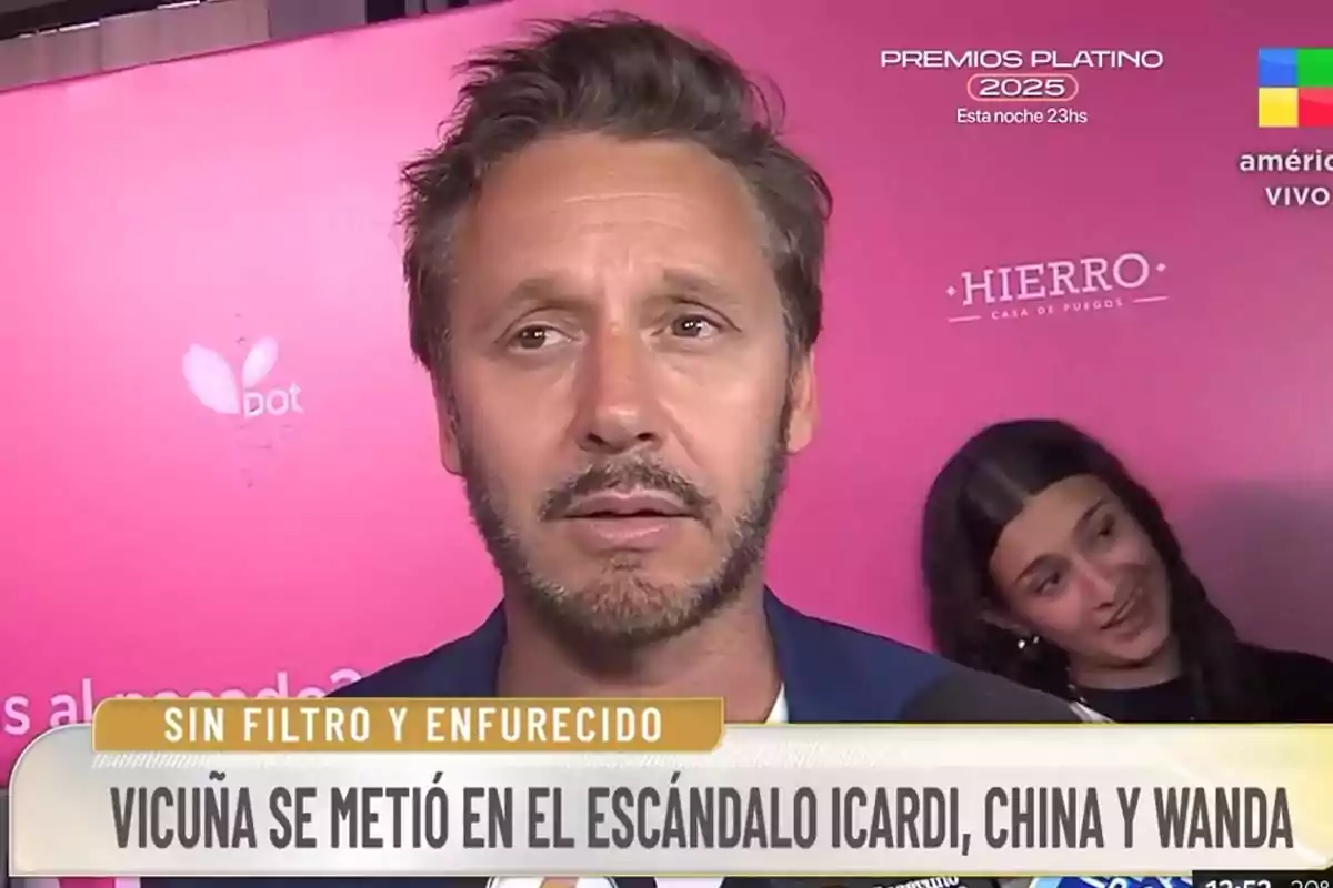 A man with a beard and short hair is being interviewed in front of a pink background, while a woman smiles behind him. A man with a beard and short hair is being interviewed in front of a pink background, while a woman smiles behind him.