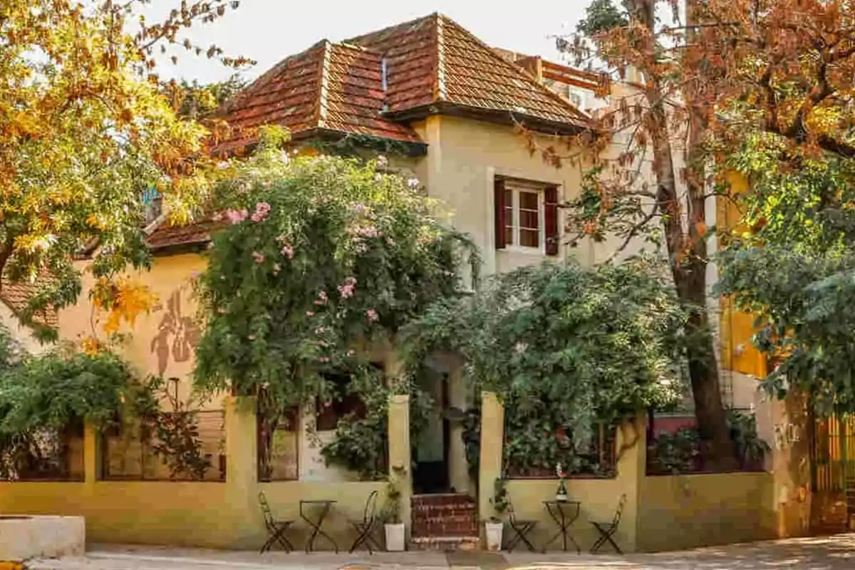 Two-story house with a red tile roof surrounded by trees and plants, with tables and chairs on the sidewalk in front of the entrance Two-story house with a red tile roof surrounded by trees and plants, with tables and chairs on the sidewalk in front of the entrance
