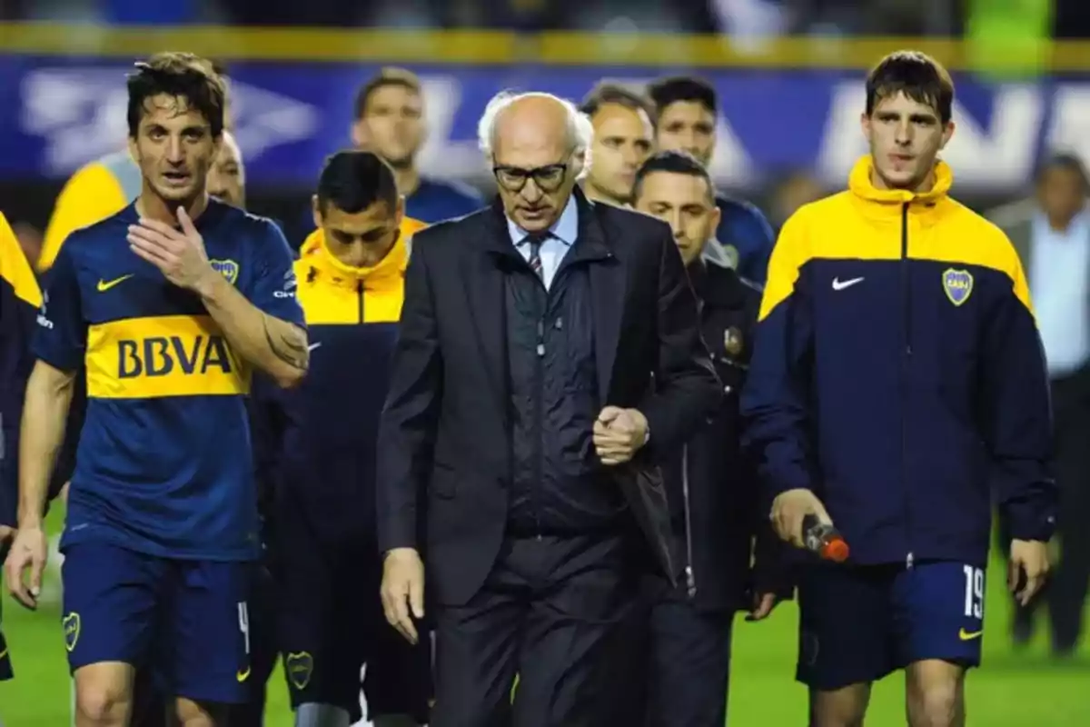 A group of people wearing Boca Juniors sportswear walks alongside an older man dressed in a suit on a soccer field. A group of people wearing Boca Juniors sportswear walks alongside an older man dressed in a suit on a soccer field.