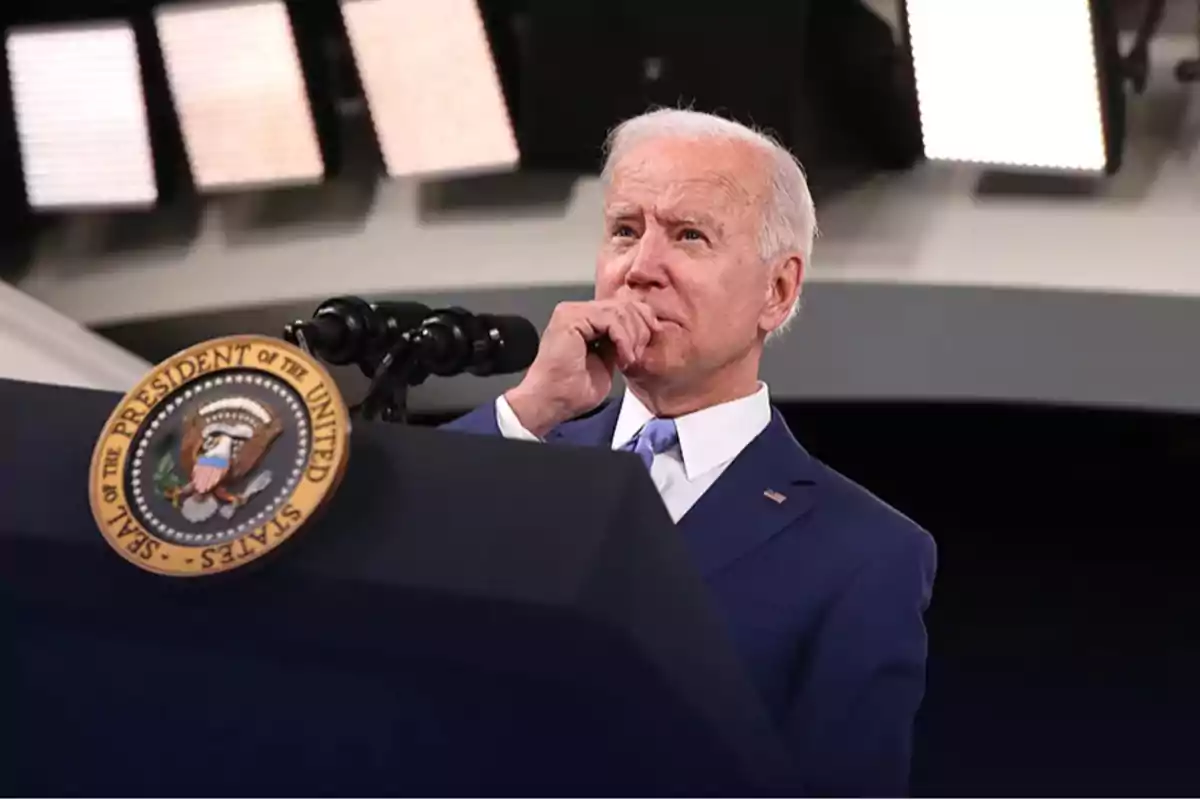 A man in a blue suit is standing behind a podium with the presidential seal of the United States, with bright lights in the background.