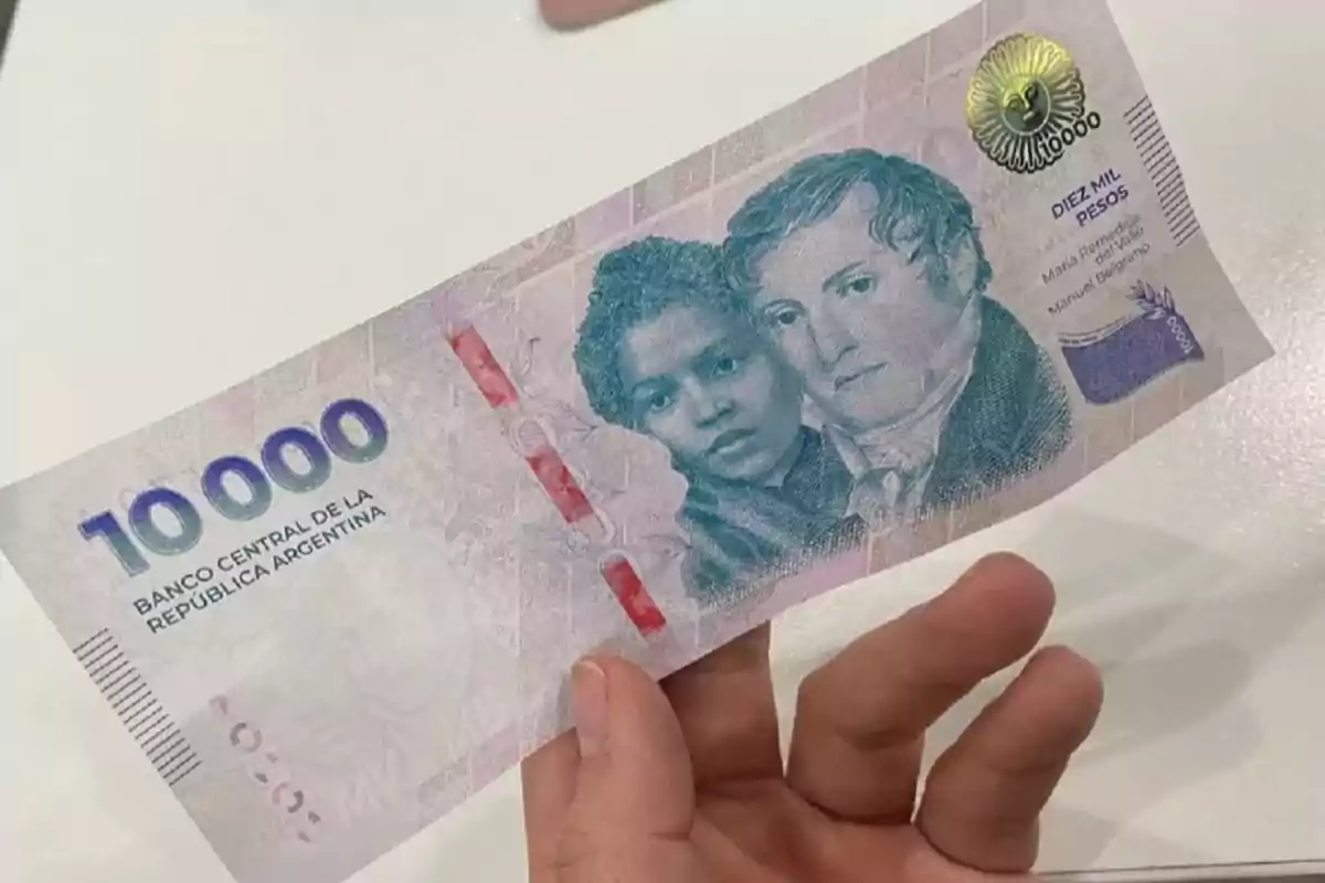 Una mano sosteniendo un billete de 10,000 pesos argentinos con imágenes de dos personas en el centro.