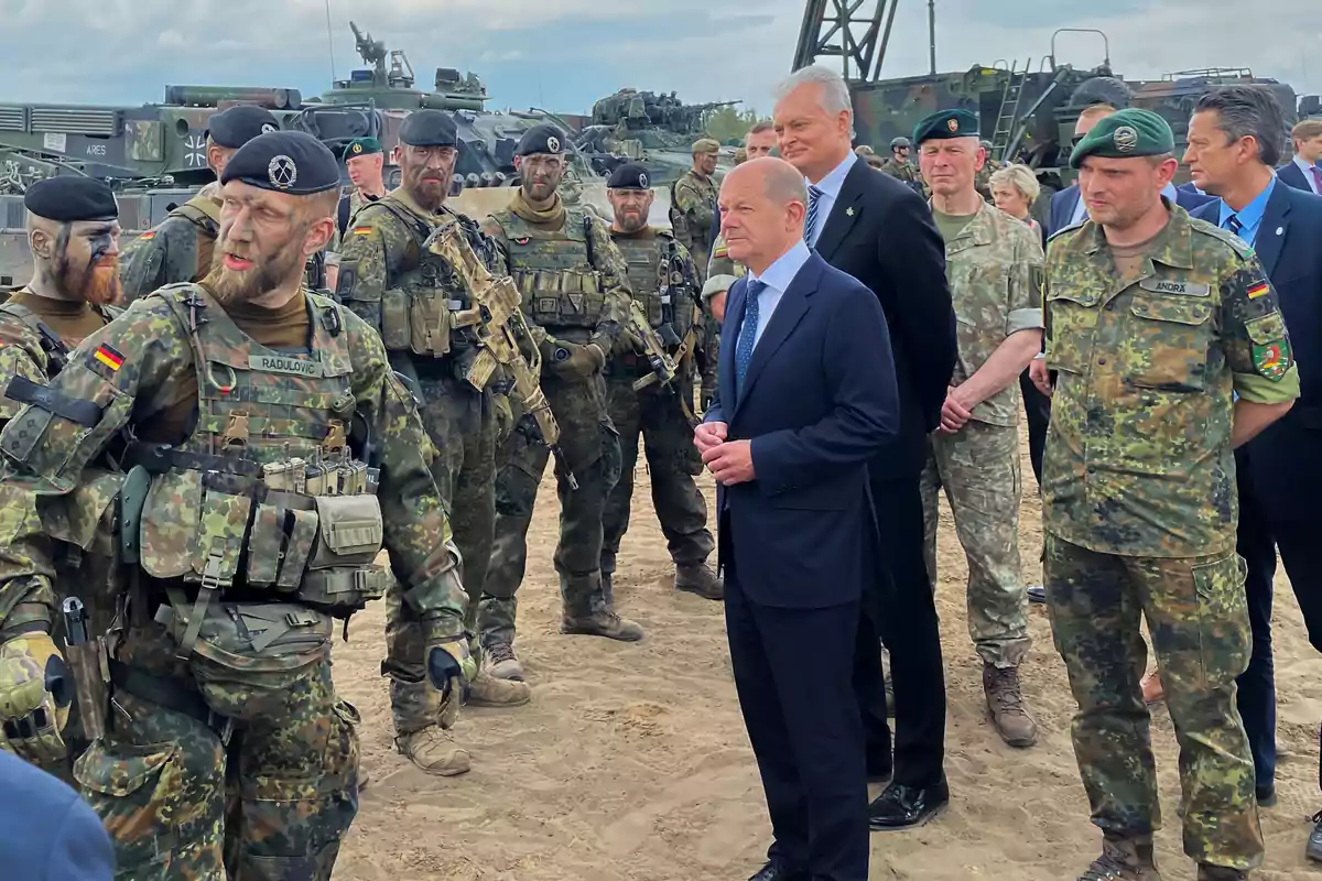 A group of military personnel in camouflage uniforms is gathered in an outdoor area alongside several men in formal suits, with military vehicles visible in the background. A group of military personnel in camouflage uniforms is gathered in an outdoor area alongside several men in formal suits, with military vehicles visible in the background.
