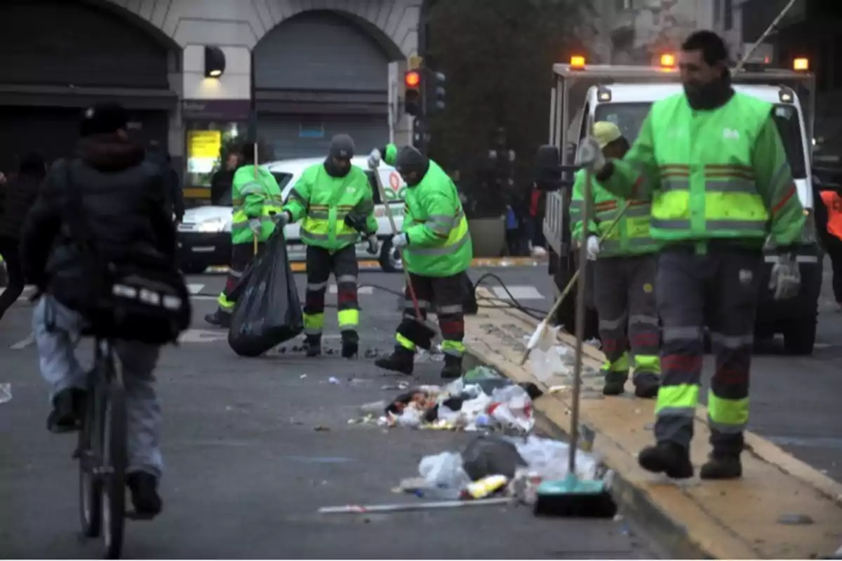 Cleaning workers in reflective vests pick up trash on an urban street as a cyclist passes by.