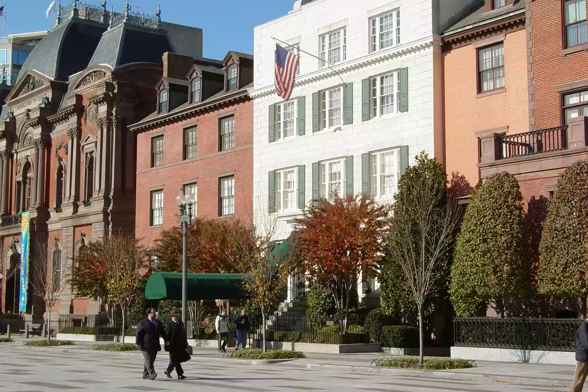 Fachada de edificios históricos con árboles y personas caminando por la acera en un día soleado Fachada de edificios históricos con árboles y personas caminando por la acera en un día soleado