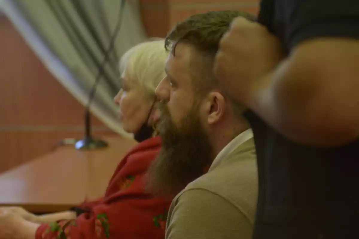 Dos personas sentadas en una sala, una de ellas con barba y la otra con cabello claro, observando hacia el frente.