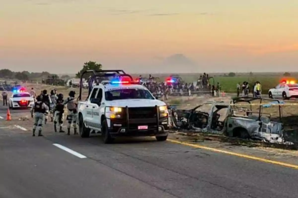 Una escena en una carretera con vehículos de la policía y militares, junto a un coche quemado, con luces de emergencia encendidas y un grupo de personas al fondo.