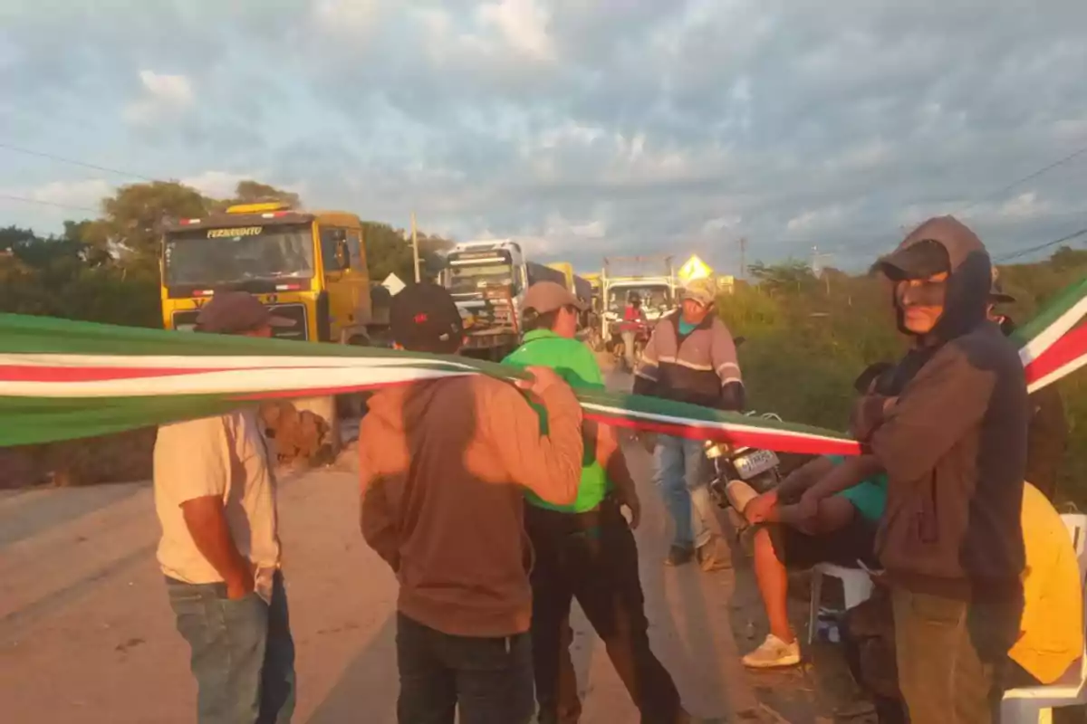Un grupo de personas sostiene una bandera en medio de una carretera bloqueada, con varios camiones detenidos al fondo bajo un cielo nublado. Un grupo de personas sostiene una bandera en medio de una carretera bloqueada, con varios camiones detenidos al fondo bajo un cielo nublado.