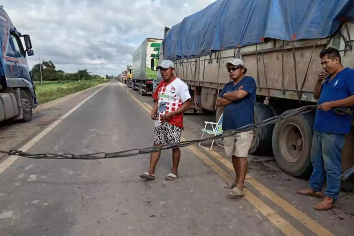 Tres hombres sostienen una cuerda bloqueando una carretera mientras varios camiones están detenidos a un lado. Tres hombres sostienen una cuerda bloqueando una carretera mientras varios camiones están detenidos a un lado.