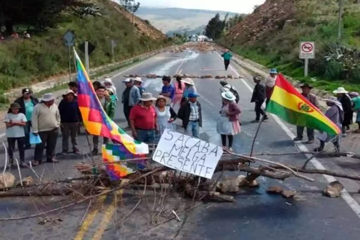 Personas bloqueando una carretera con ramas y piedras, sosteniendo banderas de Bolivia y la wiphala, y un cartel que dice Sacaba Melga Presente Personas bloqueando una carretera con ramas y piedras, sosteniendo banderas de Bolivia y la wiphala, y un cartel que dice Sacaba Melga Presente