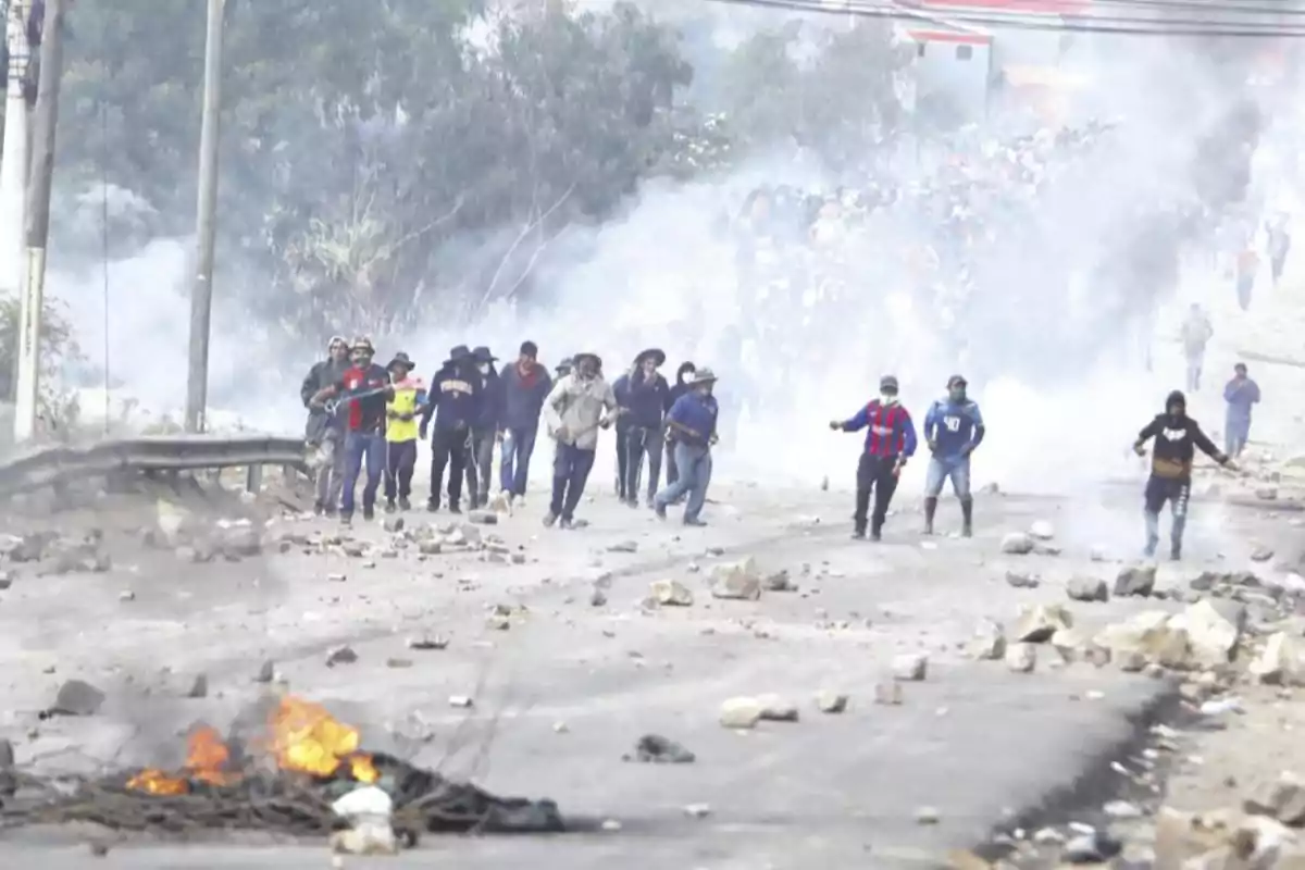 Un grupo de personas avanza por una calle llena de piedras y humo mientras una barricada arde en primer plano