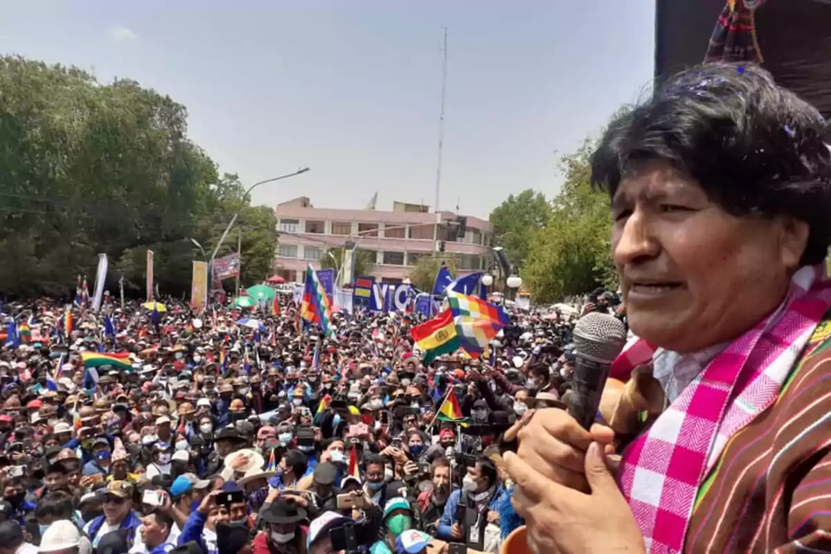 Una persona hablando en un mitin al aire libre con una multitud de personas sosteniendo banderas coloridas.