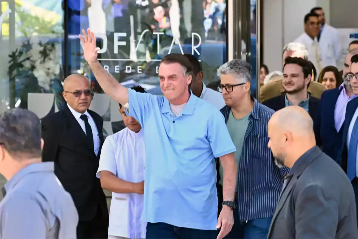 A man in a blue jersey waves while walking surrounded by several people in front of a building.