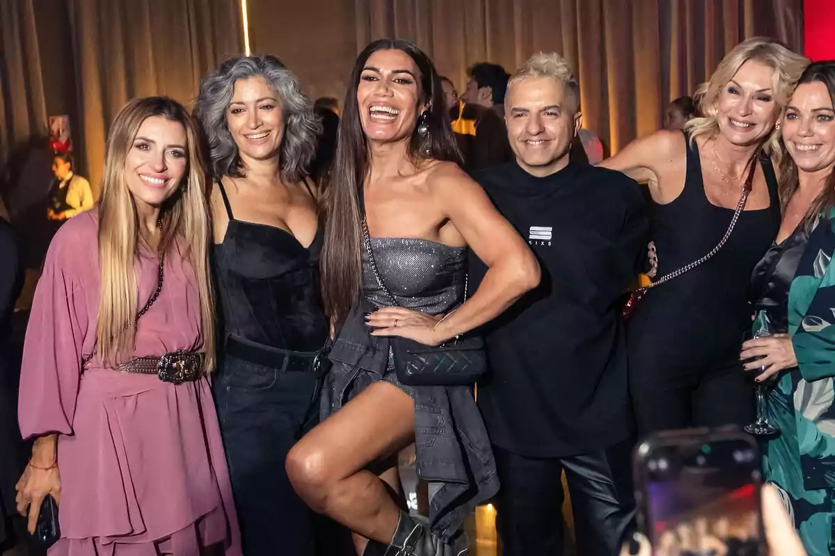 Un grupo de personas sonríe y posa para una foto en un evento social, con cortinas de fondo y luces cálidas. Un grupo de personas sonríe y posa para una foto en un evento social, con cortinas de fondo y luces cálidas.