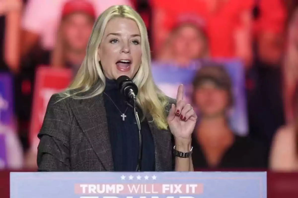 Una mujer rubia hablando en un mitin político con un micrófono frente a un cartel que dice 