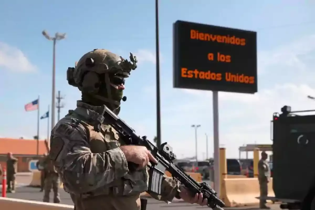 Un soldado armado con uniforme militar y equipo táctico está de pie frente a un cartel que dice "Bienvenidos a los Estados Unidos" en un puesto fronterizo.