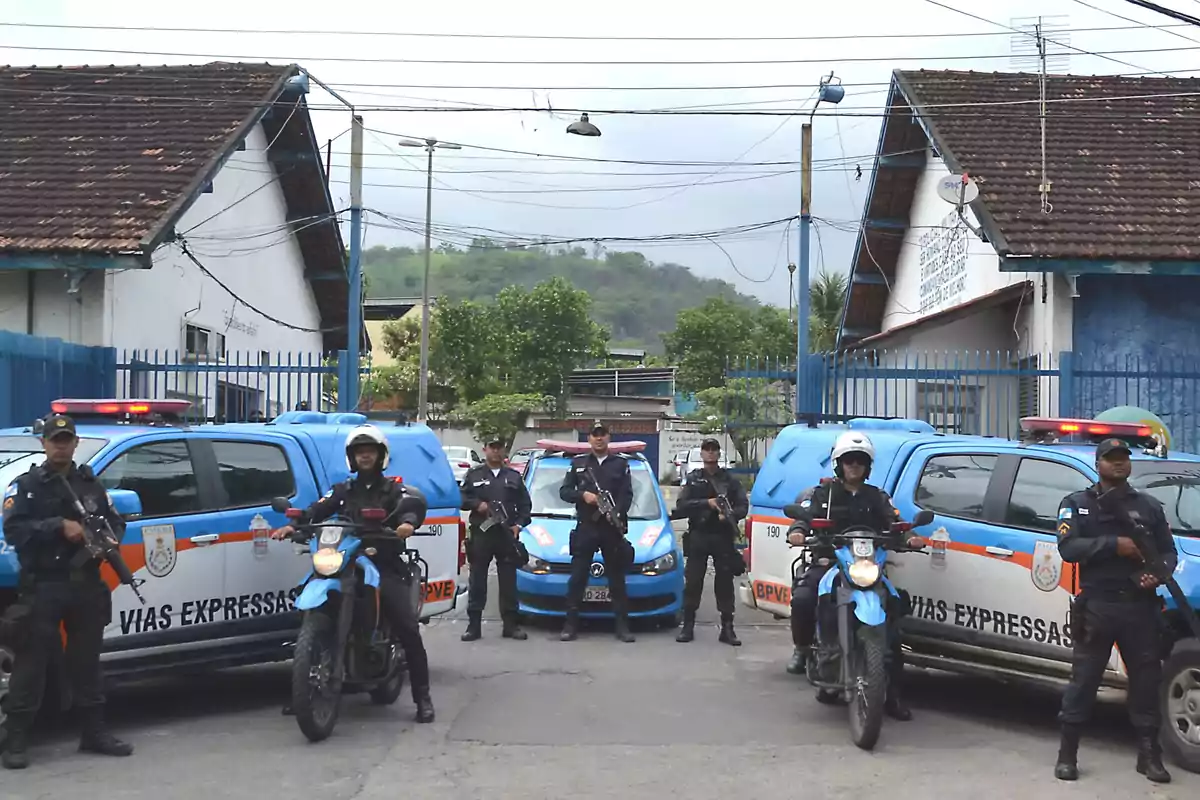 Un grupo de policías armados está de pie frente a vehículos policiales, incluyendo motocicletas y camionetas, en una calle entre dos edificios con techos de tejas. Un grupo de policías armados está de pie frente a vehículos policiales, incluyendo motocicletas y camionetas, en una calle entre dos edificios con techos de tejas.