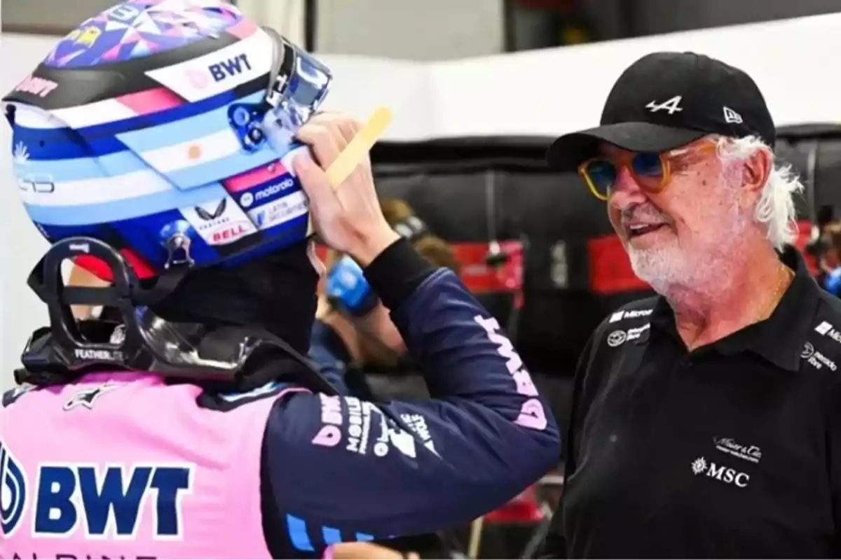 A racing driver wearing a helmet and BWT Alpine uniform talks with an older man wearing sunglasses and a black cap. A racing driver wearing a helmet and BWT Alpine uniform talks with an older man wearing sunglasses and a black cap.
