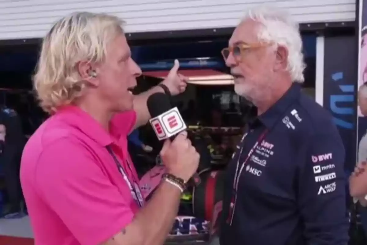Un reportero con camisa rosa entrevista a un hombre mayor con barba blanca y gafas, ambos están frente a un garaje de automovilismo. Un reportero con camisa rosa entrevista a un hombre mayor con barba blanca y gafas, ambos están frente a un garaje de automovilismo.
