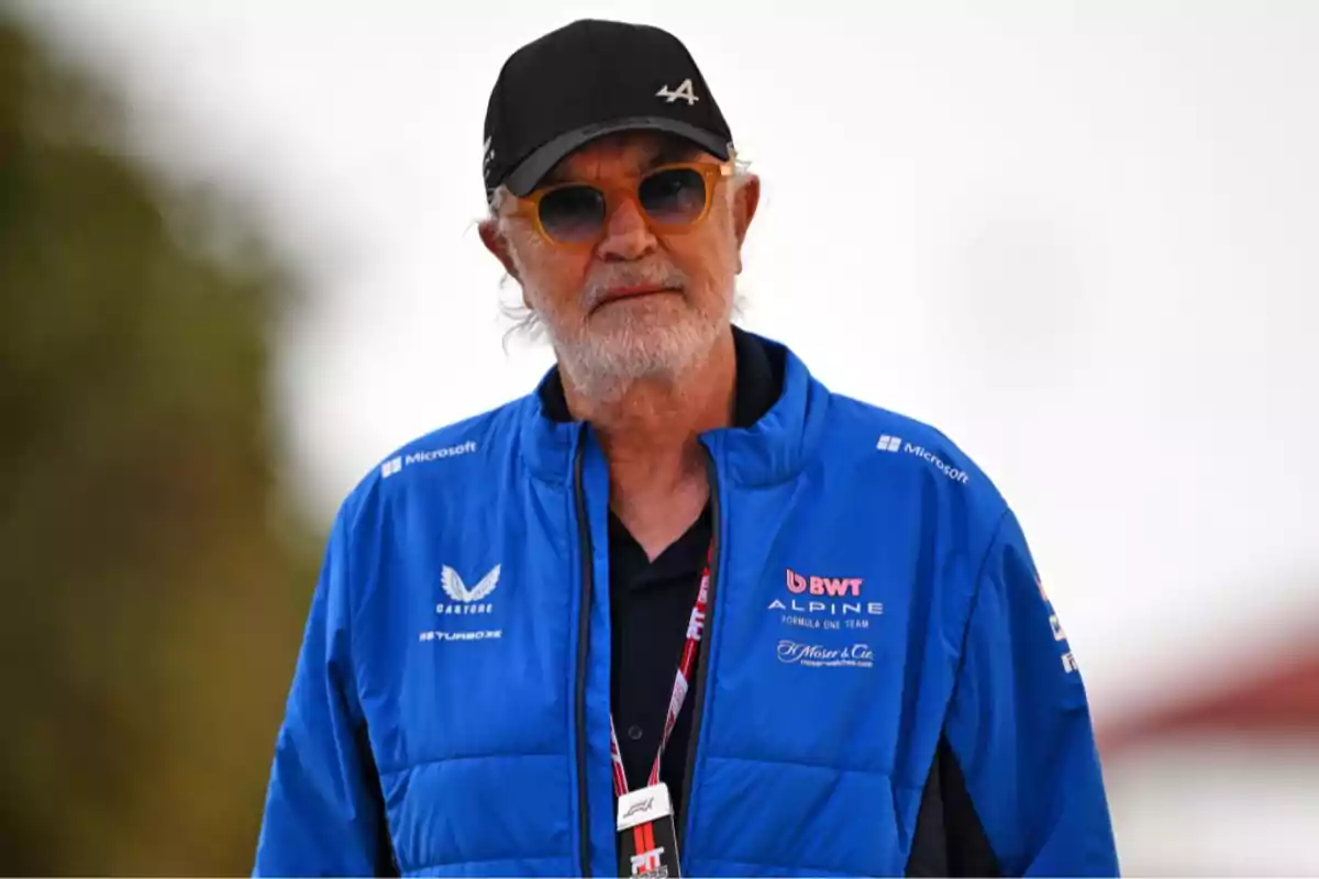 Hombre mayor con barba y gafas de sol vistiendo una gorra negra y una chaqueta azul del equipo Alpine de Fórmula 1