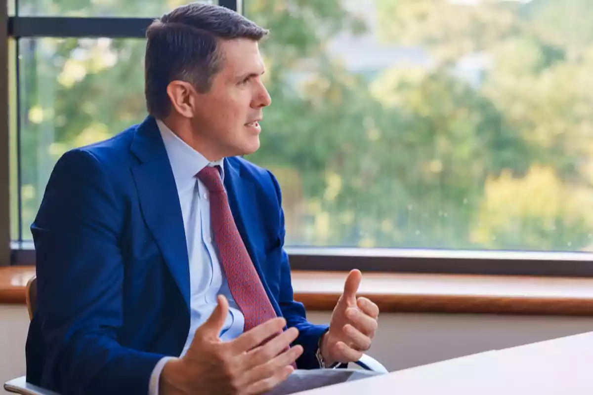 A man in a blue suit and red tie gestures while sitting next to a window overlooking a green landscape. A man in a blue suit and red tie gestures while sitting next to a window overlooking a green landscape.