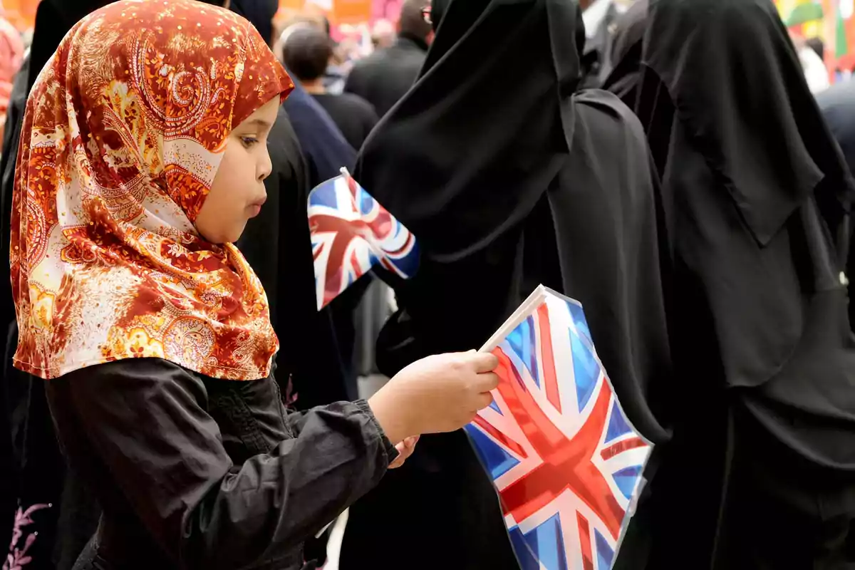 Una niña con un pañuelo colorido sostiene una bandera del Reino Unido en un evento concurrido.