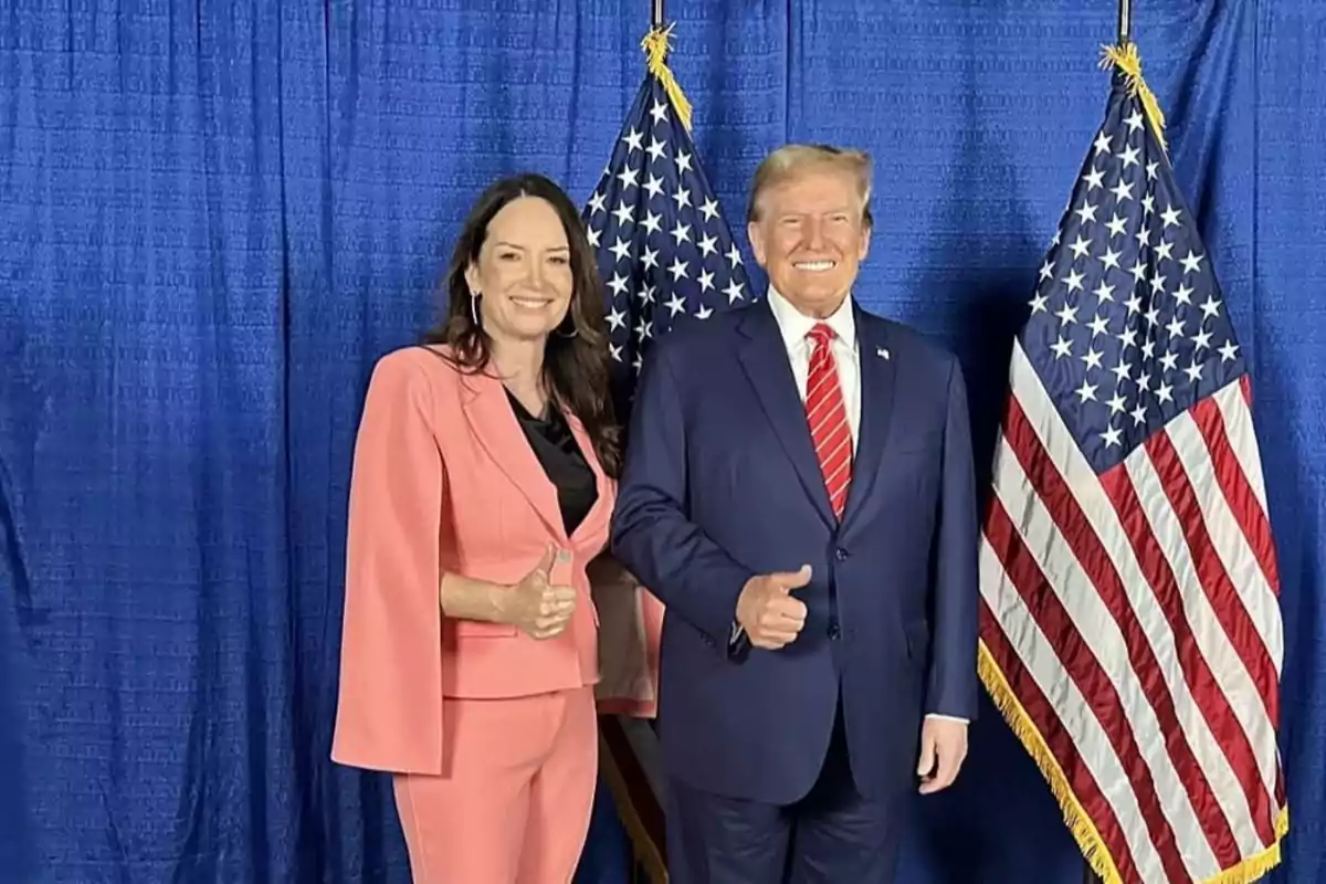 A woman in a pink suit and a man in a blue suit pose smiling in front of two American flags and a blue background, both giving a thumbs-up gesture. A woman in a pink suit and a man in a blue suit pose smiling in front of two American flags and a blue background, both giving a thumbs-up gesture.