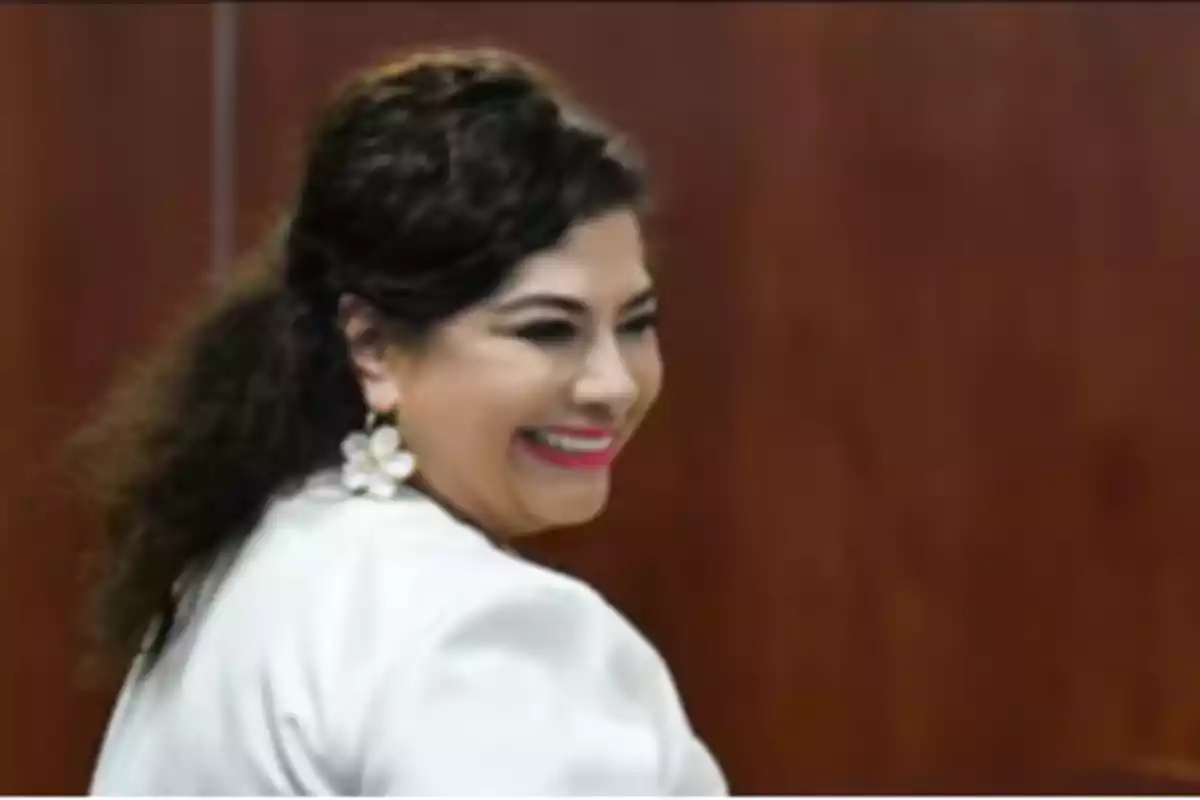 Una mujer sonriente con cabello oscuro y aretes de flor, vestida de blanco, frente a un fondo marrón.