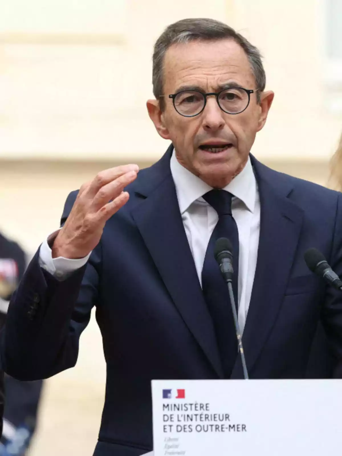 Hombre de mediana edad con gafas y traje oscuro hablando en una conferencia de prensa frente a un atril del Ministerio del Interior y de Ultramar de Francia Hombre de mediana edad con gafas y traje oscuro hablando en una conferencia de prensa frente a un atril del Ministerio del Interior y de Ultramar de Francia