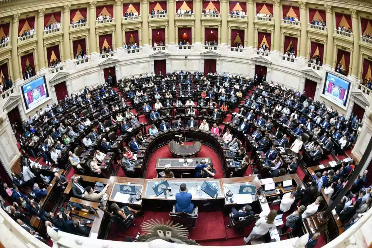 Una vista panorámica de un parlamento lleno de personas sentadas en sus escaños, con una arquitectura interior elegante y pantallas en las paredes.