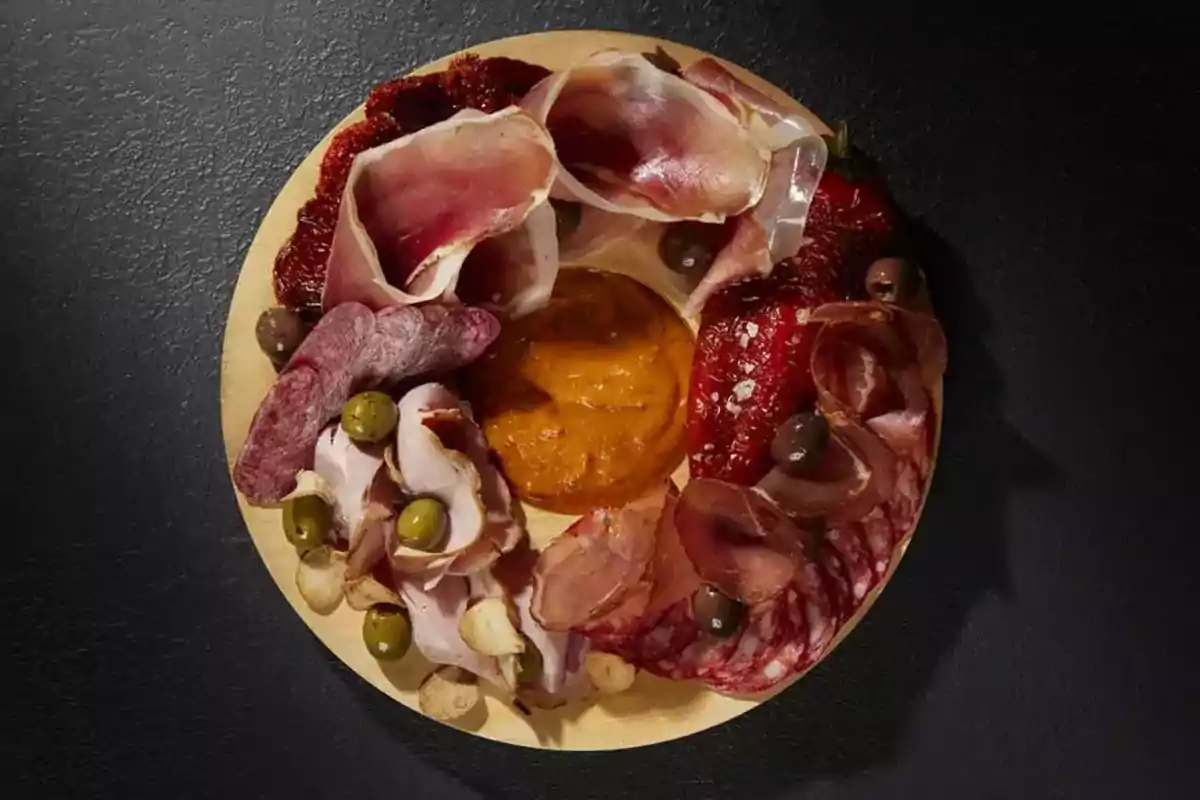 Assorted cold cuts platter with olives and a dip in the center on a dark surface Assorted cold cuts platter with olives and a dip in the center on a dark surface