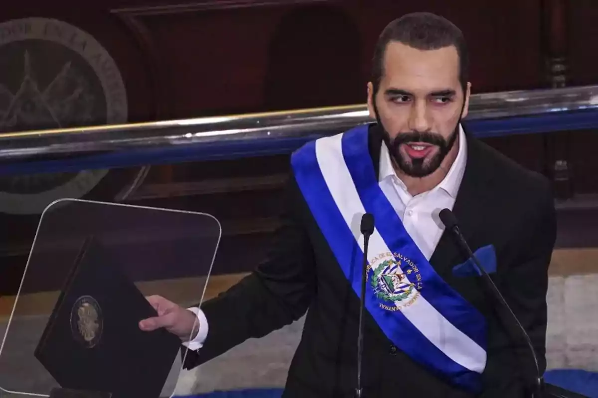 A man with a beard and short hair wears a blue and white presidential sash while holding a document and speaking into a microphone in a formal setting. A man with a beard and short hair wears a blue and white presidential sash while holding a document and speaking into a microphone in a formal setting.