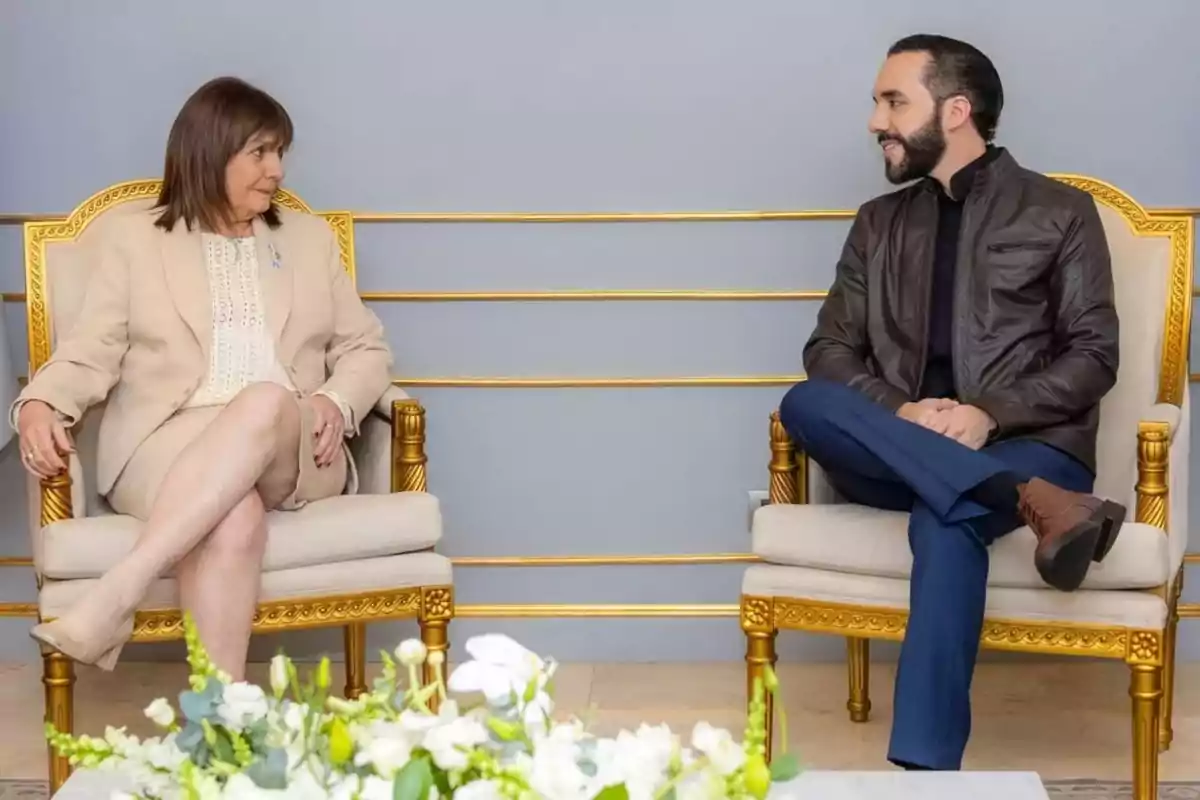 Dos personas sentadas en sillas doradas conversan en una sala decorada con flores blancas. Dos personas sentadas en sillas doradas conversan en una sala decorada con flores blancas.
