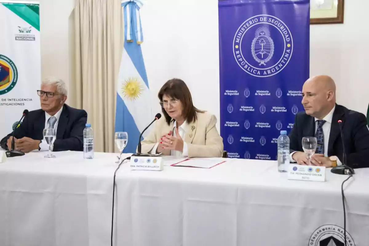 Tres personas sentadas en una mesa durante una conferencia de prensa con banderas y carteles del Ministerio de Seguridad de Argentina en el fondo.