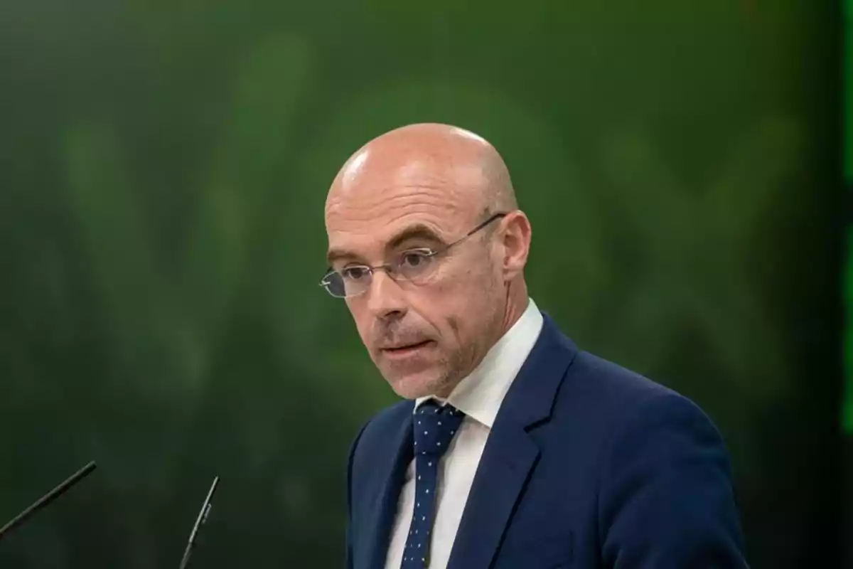 Un hombre calvo con gafas y traje azul oscuro habla frente a un micrófono con un fondo verde.